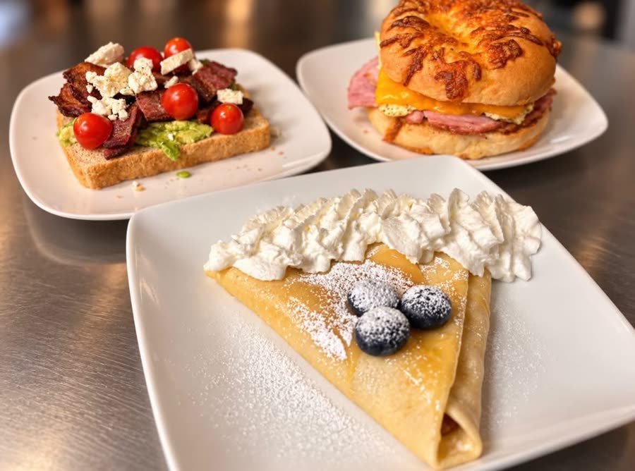 A folded crepe dusted with powdered sugar, topped with whipped cream and three blueberries, with avocado toast and a ham and cheese bagel sandwich behind it