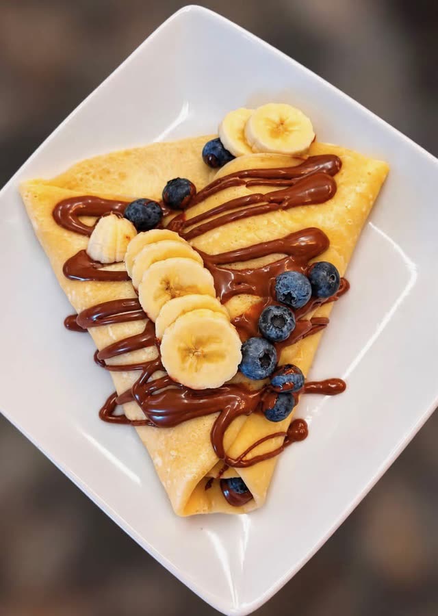 A folded crepe on a white plate, seen from above, with chocolate drizzle, banana slices and blueberries