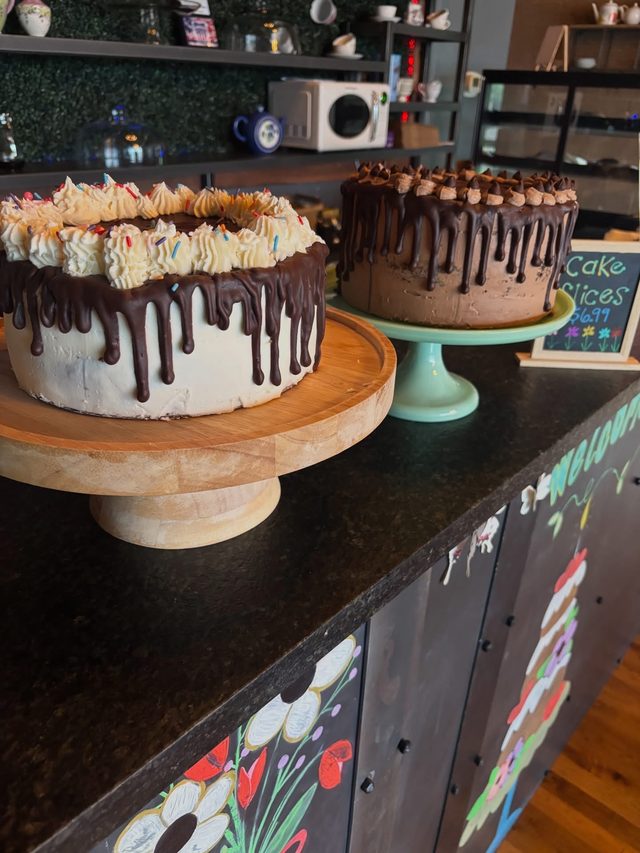 Two chocolate-drip cakes on a wooden stand and a mint-green stand on the counter, beside a chalkboard sign for cake slices